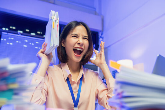 Stressed young Asian businesswoman overwhelmed with workload and holding documents while screaming in office.