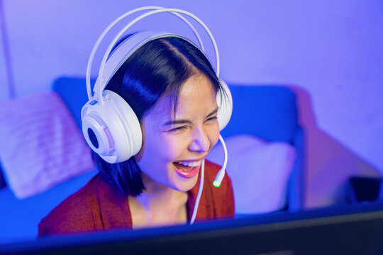 Excited young Asian girl playing online computer game with headset and microphone at home, esports and live streaming concept.