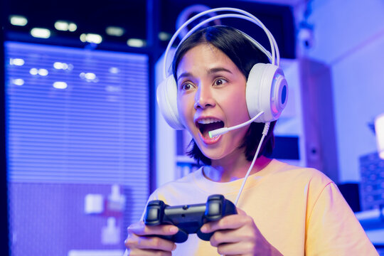 Excited young Asian girl playing video game with gaming headset and controller at home, esports online concept.