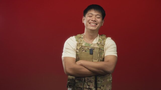 Young man in camouflage tactical vest and dog tags, smiling with arms crossed in studio against a red backdrop; pride camaraderie.