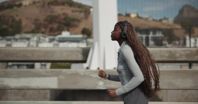 Running, headphones and black woman in city for fitness, cardio health and endurance training. Workout, jog and person streaming music for exercise, wellness and listening to audio on bridge outdoor