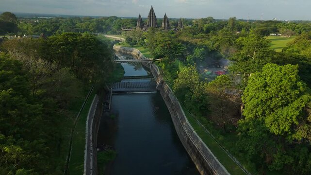 Aerial view of a narrow canal flowing through lush green forests with the ancient Prambanan Temple visible in the distance Kecamatan Prambanan, Daerah Istimewa Yogya, Indonesia.