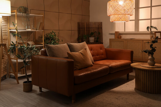 Interior of dark living room with brown sofa, plants and glowing lamps at night