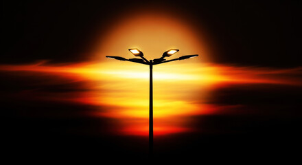 Fototapeta premium A tall street light stands silhouetted against a vibrant, fiery sunset. Two light fixtures illuminate the darkening sky, casting a warm glow.