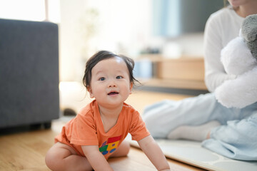Fototapeta premium Cute toddler smiles brightly on wooden floor. Baby wears orange shirt, eyes wide with joy. Parent sits nearby, holding soft toy. Soft natural light fills cozy, warm room. Perfect for parenting, family