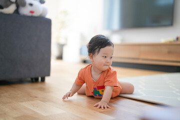 Naklejka premium Cute baby sits on wooden floor. Child wears orange shirt with colorful patch. Baby looks off-camera, showing quiet curiosity. Room has soft light and cozy furnishings. Image suits parenting