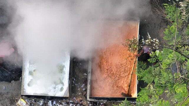 Natore, Bangladesh - 13 March 2024: Aerial view of traditional jaggery production showing workers stirring boiling molasses in large vats with thick steam rising.