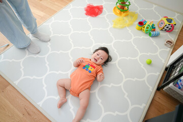 Naklejka premium Baby lies smiling on soft patterned mat. Tiny hands reach toward colorful toys nearby. Adult feet stand close, watching calmly. Bright yellow ball and plush items scattered around
