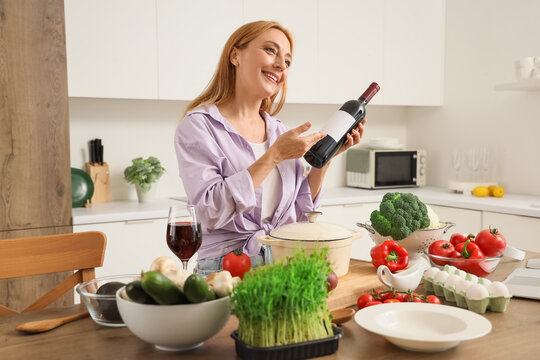Mature woman with bottle of wine recording cooking video class in kitchen