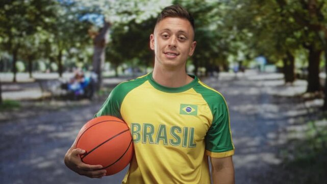 Man holding basketball with hand, wearing brasil jersey and flag patch, smiling young blond player standing on street lined with trees and benches; team pride.