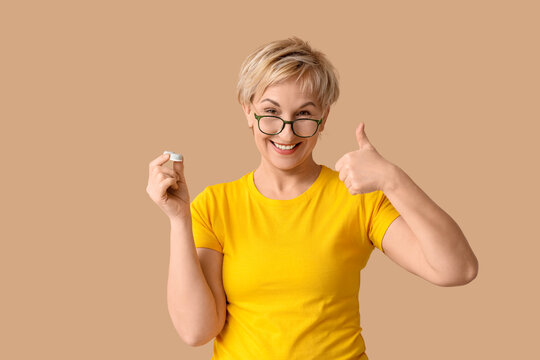 Mature woman in eyeglasses with contact lens case showing thumb-up gesture on brown background