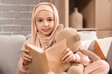 Obraz premium Little Muslim girl in hijab with teddy bear reading book at home