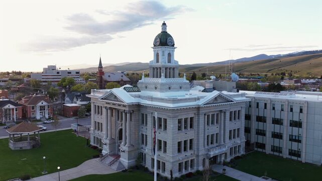 Missoula historic courthouse with white neoclassical facade and green clock dome, church steeple and brick buildings surrounding, golden rolling hills and mountain range. Montana, USA. Aerial descend.