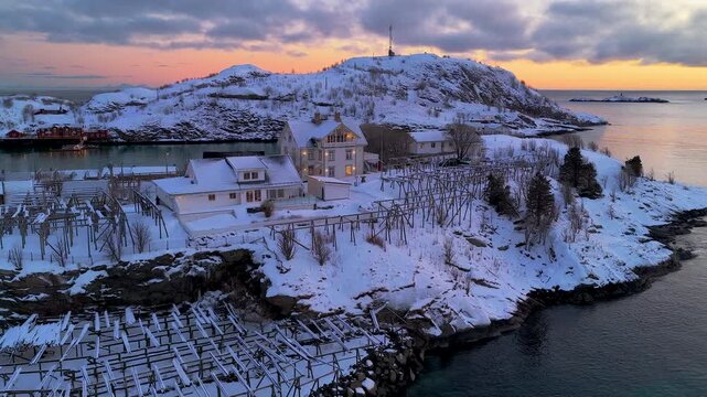 Cinematic aerial shot of a snowcovered fishing village in Lofoten, Norway during golden hour. Features traditional houses and fish drying racks against a pink sunset sky.