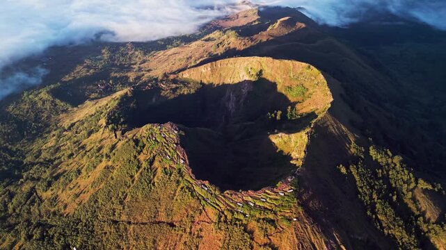 Volcanic caldera of Mount Batur in Bali with steep crater walls and rugged terrain illuminated by warm morning light with forested slopes surrounding the crater in wide aerial drone view
