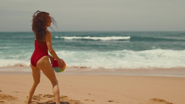 Caucasian female beachgoer in red swimsuit plays with colorful beach ball, running along sandy shore with ocean waves and another woman in bikini visible in the background
