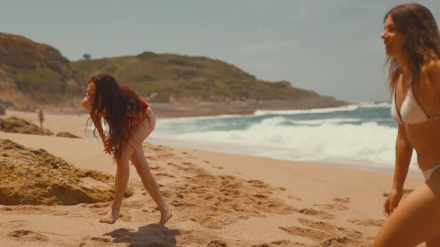 Two women playing catch with a beach ball on sandy shore, one in red swimsuit and the other in white bikini, ocean waves and green hills in the background
