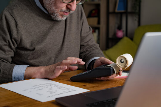 Senior man calculating expenses and checking receipt with calculator at home desk