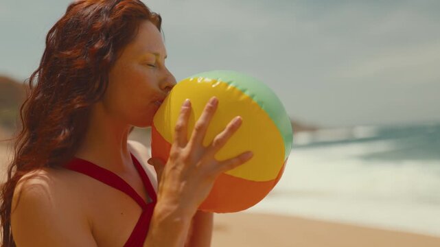 Woman with long red hair inflates a colorful beach ball at the shoreline, with gentle waves and sandy beach creating a vibrant summer atmosphere