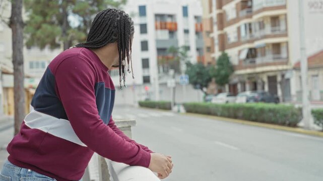 Young man leans on concrete balustrade with clasped hands, braided hair and sweatshirt, looking down an empty street; contemplation calm.