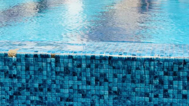 Close up of luxury infinity swimming pool edge with blue mosaic tiles and crystal clear water ripple reflections. Premium resort spa wellness area, aquatic texture and hotel design concept.