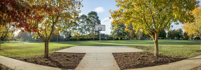 Obraz premium Panoramic view of an outdoor half basketball court framed by autumn trees in spacious municipal suburban park. Council recreational facility, civic amenities, family-friendly neighborhood in Australia