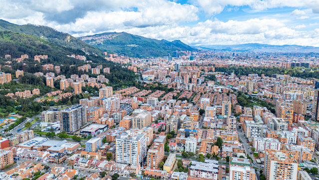 Vast cityscape of Bogot&aacute; showcasing high-rise buildings and residential areas nestled against a mountainous backdrop under a partly cloudy sky.