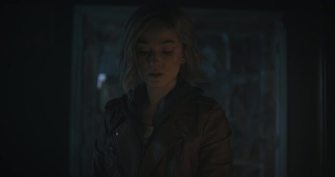 Young blonde woman walking through dark room with raging storm outside as she stares down at something