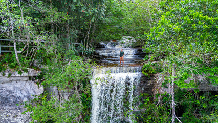 A lone individual stands on rocks beside a cascading waterfall, surrounded by dense greenery and tropical plants in Colombia. © Jhampier Giron