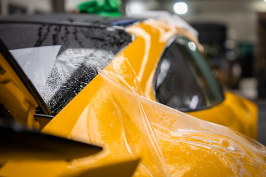 Close view of a yellow luxury sports car as clear film is applied on a wet soapy panel, showing water beads, squeegee marks, and a lifted edge under soft indoor light.
