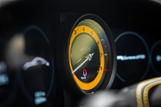 Driver seat view of Porsche 911 Carrera GTS cluster, GTS on tachometer, 0 kmh shown. Needle rests at idle, soft bokeh, yellow stitching, low ambient light.
