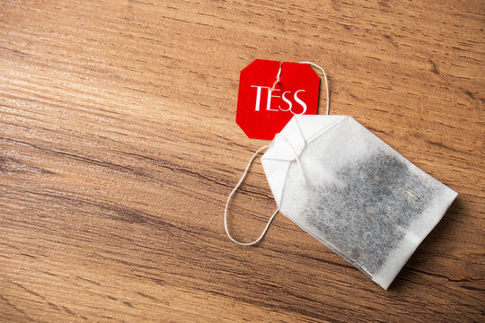 A Tess brand tea bag with its red tag rests on a wooden surface. The tea bag contains dark tea leaves.