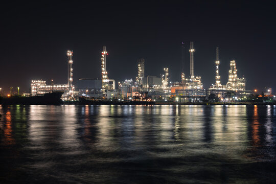 Oil refinery plant at night in Bangkok, Thailand, industrial complex glowing beneath dark sky. Lights reflect across rippling water, highlighting processing towers, pipelines and energy infrastructure