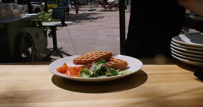 Outdoor brunch. Culinary leisure. Unrecognizable skilled chef man hands pouring hollandaise sauce on egg benedict cooking waffle salad salmon dish on plate.