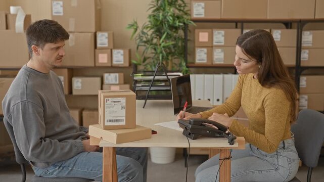 Man and woman sign parcel forms and exchange a pen over stacked boxes in a building; small business teamwork trust.