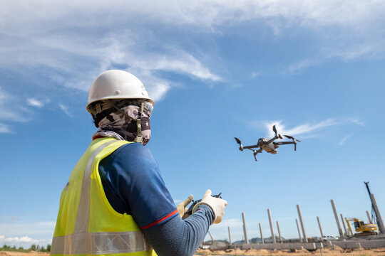 Drone Surveying at Construction Site: A construction worker in a hard hat and safety vest operates a drone using a remote control, surveying an active building site under a clear blue sky.