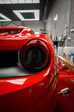 Three quarter view shows circular taillight and sculpted bodywork. Industrial gray brick wall, grid lighting, red mats, tools, and glossy reflections suggest precision.