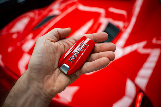 A person holds a glossy red Ferrari key fob, chrome Ferrari script visible. Blurred red 488 Spider bodywork and vents sit parked indoors under bright reflective light.