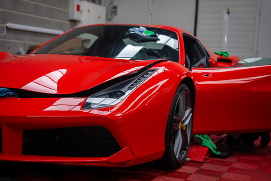 Close up low angle view of a red Ferrari 488 Spider in a detailing garage, with LED headlight, five spoke wheel, yellow caliper, open door, and towels on red mat.