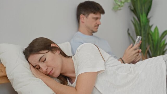 Woman lying on pillow on bed under white bedding while man sits against headboard and scrolls smartphone in a building; quiet loneliness.