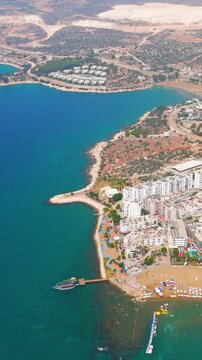 Vertical video. Kizkalesi, Mersin, Turkey. Aerial view of Mediterranean coastal resort town with dense white hotel buildings, turquoise bay, and sandy beach under clear summer sky.. Aerial View. Rich