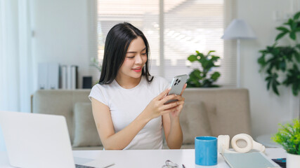 Fototapeta premium Young Asian woman sitting at desk, smiling and looking at smartphone, enjoying connectivity and digital lifestyle