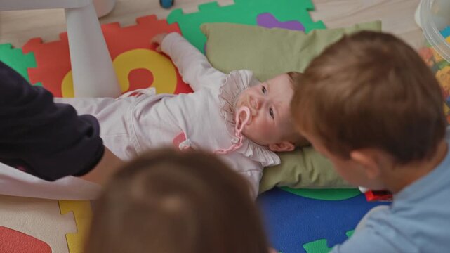 Toddler girl crying with pacifier and outstretched hand on colorful foam play mat inside building; distress.