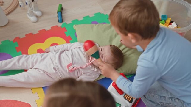 Toddler boy kneels beside infant girl lying with pacifier on colorful foam play mat, holding toy cash register and gently reaching toward her in building; tender moment.