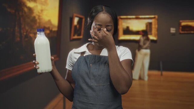 Woman wearing apron holds milk bottle and pinches nose, eyes closed, in an art gallery building with framed paintings; disgust reaction.