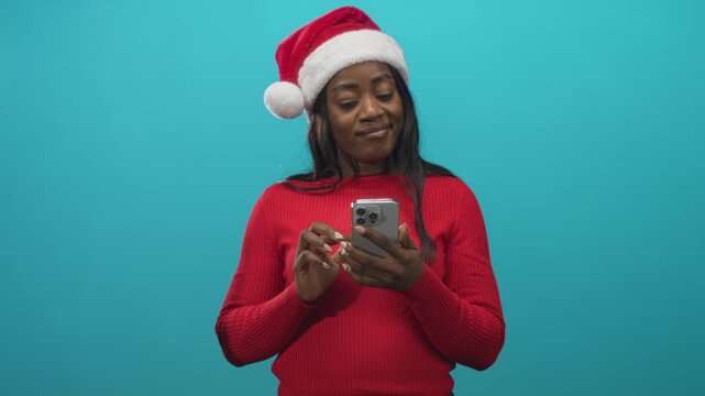 Woman in santa hat and red sweater holds smartphone, typing with fingers and smiling while looking at screen in studio; holiday cheer.