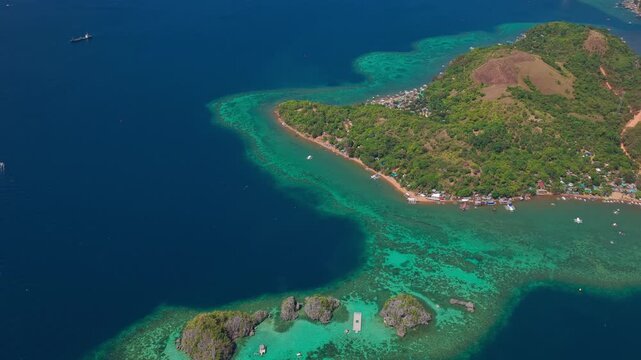 Aerial backwards reveal shot pulling away from a lush green island headland and coral reef in Coron, Palawan.