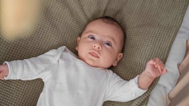 Baby in cradle on textured olive blanket raises arms and clenches fist showing grasp reflex while lying in a residential building nursery; calm curiosity.