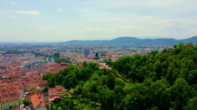 Graz, Austria. The Uhrturm clock tower with reversed hands, perched on Schlossberg hill above dense terracotta rooftops, aerial panoramic view under bright summer sky. Drone footage