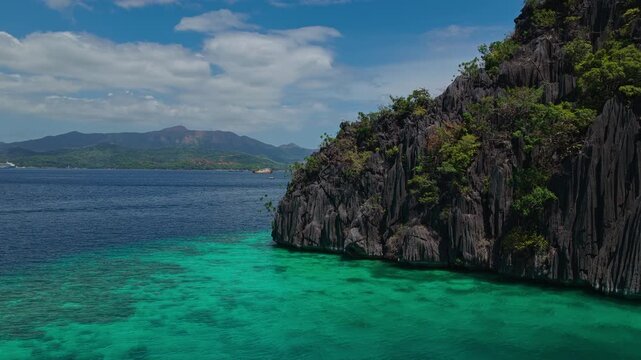 Drone orbit shot around a dramatic limestone headland cliff surrounded by turquoise ocean in Coron, Philippines.